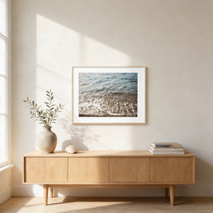 a serene indoor setting with a wooden cabinet, a framed picture of a beach scene, a vase with greenery, and a stack of books.