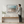 A man is standing in front of a large framed picture of a beach, looking at it intently. The picture depicts a serene beach scene with waves gently lapping against the shore. The man appears to be admiring the artwork, which is mounted on a wall above a wooden bench.